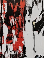 A chaotic and abstract composition featuring bold, distressed patterns in black, red, and white, with overlapping layers and vertical streaks. The forms suggest the remnants or peeling layers of street art or graffiti.