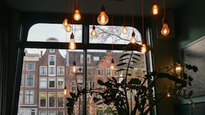 A cozy indoor setting with several warm light bulbs hanging from the ceiling in front of a large window. The view through the window shows traditional European-style buildings with brick facades. A silhouetted plant is visible in the foreground, adding to the intimate atmosphere.