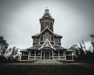 A tall, wooden church with a steeple stands against an overcast sky. The structure is painted in red with white trims, and has a symmetric design featuring large windows and a prominent entrance. Surrounding the church are sparse trees.