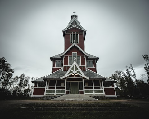 A tall, wooden church with a steeple stands against an overcast sky. The structure is painted in red with white trims, and has a symmetric design featuring large windows and a prominent entrance. Surrounding the church are sparse trees.