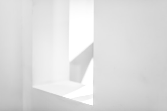 A minimalist white space featuring geometric shapes and clean lines. There is a rectangular opening on the right side creating shadows and light contrasts.