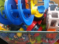 A metal wire basket filled with a variety of colorful children's toys including toy cars, construction vehicles, and other plastic toys. The toys are primarily blue, yellow, red, and green.