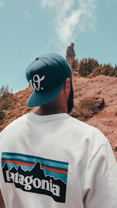 A person wearing a white 'Patagonia' shirt and a blue cap stands with their back to the camera, looking at a rocky landscape with sparse vegetation and a distinctive rock formation under a clear sky.