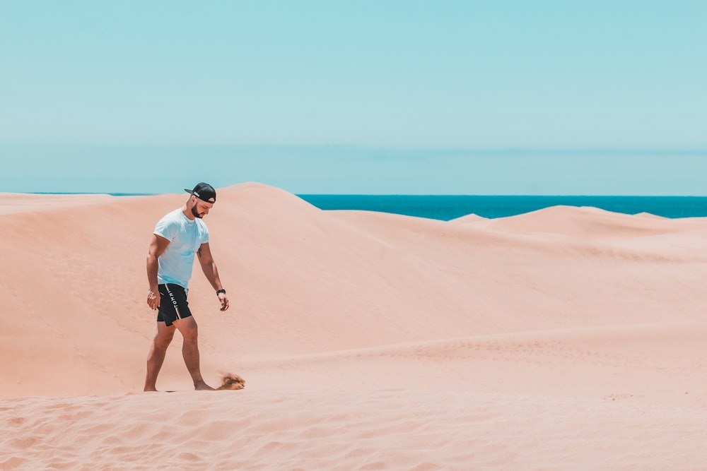 ティール色のクルーネックシャツを着て、砂丘を歩く黒いショートパンツを着た男性