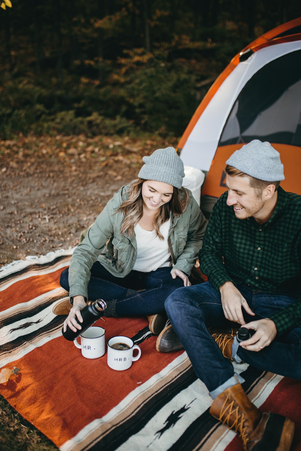 smiling man and woman sitting outside tent with two mugs