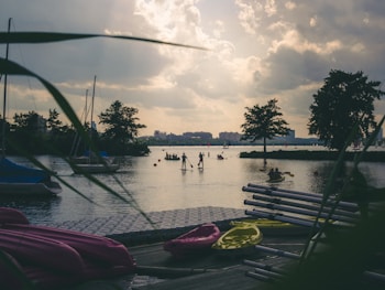 A scenic lakeside view featuring people engaging in paddleboarding and kayaking. Several boats and kayaks are visible on the dock in the foreground, with lush greenery framing the image. The calm water reflects the cloudy yet bright sky, and trees line the opposite shore.