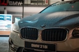 A close-up view of a BMW 218i car parked outdoors, with noticeable water droplets on its hood, reflecting the ambient lights. The dealership is visible in the background, featuring a modern, illuminated showroom.