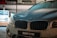 A close-up view of a BMW 218i car parked outdoors, with noticeable water droplets on its hood, reflecting the ambient lights. The dealership is visible in the background, featuring a modern, illuminated showroom.