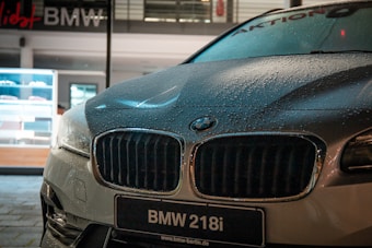 A close-up view of a BMW 218i car parked outdoors, with noticeable water droplets on its hood, reflecting the ambient lights. The dealership is visible in the background, featuring a modern, illuminated showroom.
