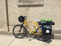 A yellow bicycle is parked against a beige brick wall. The bicycle is fully equipped with touring gear, including a black front bag, water bottles, a sleeping bag, and other camping equipment secured on the rear rack. Panniers are attached to the sides of the bike.