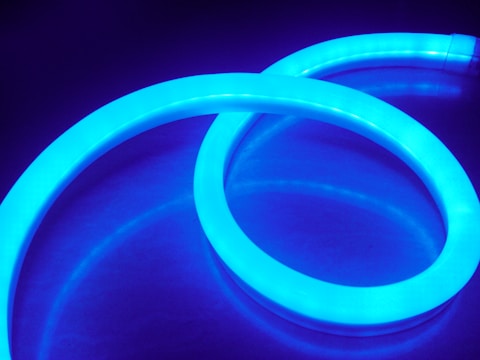 A glowing, flexible neon tube emits a vibrant blue light, creating an abstract and futuristic visual against a dark background. The tube is coiled in a smooth, curving pattern.