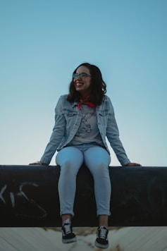 smiling woman wearing blue denim jacket sitting on black concrete surface during daytime