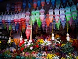Colorful hanging decorations and artificial flowers fill the space, creating a vibrant, festive atmosphere. The decorations feature intricate designs in a variety of bright colors and are suspended above a multitude of artificial flowers arranged below.