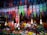 Colorful hanging decorations and artificial flowers fill the space, creating a vibrant, festive atmosphere. The decorations feature intricate designs in a variety of bright colors and are suspended above a multitude of artificial flowers arranged below.
