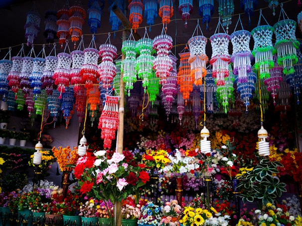 Colorful hanging decorations and artificial flowers fill the space, creating a vibrant, festive atmosphere. The decorations feature intricate designs in a variety of bright colors and are suspended above a multitude of artificial flowers arranged below.
