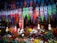 Colorful hanging decorations and artificial flowers fill the space, creating a vibrant, festive atmosphere. The decorations feature intricate designs in a variety of bright colors and are suspended above a multitude of artificial flowers arranged below.