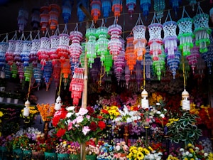 Colorful hanging decorations and artificial flowers fill the space, creating a vibrant, festive atmosphere. The decorations feature intricate designs in a variety of bright colors and are suspended above a multitude of artificial flowers arranged below.