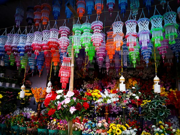 Colorful hanging decorations and artificial flowers fill the space, creating a vibrant, festive atmosphere. The decorations feature intricate designs in a variety of bright colors and are suspended above a multitude of artificial flowers arranged below.