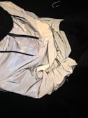 A reflective jacket is displayed with its inside lining exposed. The outer surface has a shiny, metallic appearance, while the lining features a dotted pattern. The jacket includes visible zippers and appears crumpled and spread out against a dark background.