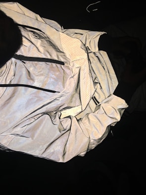 A reflective jacket is displayed with its inside lining exposed. The outer surface has a shiny, metallic appearance, while the lining features a dotted pattern. The jacket includes visible zippers and appears crumpled and spread out against a dark background.