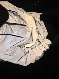 A reflective jacket is displayed with its inside lining exposed. The outer surface has a shiny, metallic appearance, while the lining features a dotted pattern. The jacket includes visible zippers and appears crumpled and spread out against a dark background.