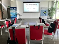 A conference room set up for a presentation or workshop with red chairs arranged around tables. Each table contains glasses, notepads, and black bags with logos. A projector screen at the front displays a title. Banners with graphics and text are positioned on either side of the room. A plant adds greenery to the setting.