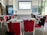 A conference room set up for a presentation or workshop with red chairs arranged around tables. Each table contains glasses, notepads, and black bags with logos. A projector screen at the front displays a title. Banners with graphics and text are positioned on either side of the room. A plant adds greenery to the setting.