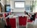 A conference room set up for a presentation or workshop with red chairs arranged around tables. Each table contains glasses, notepads, and black bags with logos. A projector screen at the front displays a title. Banners with graphics and text are positioned on either side of the room. A plant adds greenery to the setting.