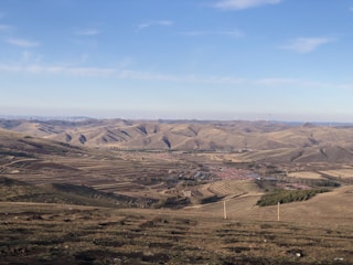 A vast landscape of rolling hills and expansive fields under a clear blue sky. The fields appear divided into neat, striped patches of farmland, suggesting agricultural activity. Small clusters of structures, possibly houses or barns, are visible in the distance. Wind turbines can be seen scattered across the hilltops, indicating a focus on renewable energy.