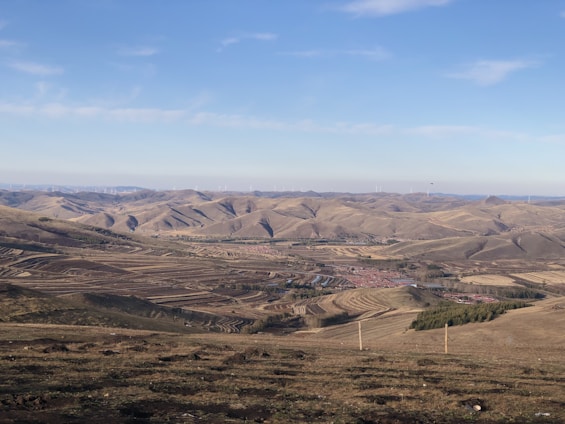 A vast landscape of rolling hills and expansive fields under a clear blue sky. The fields appear divided into neat, striped patches of farmland, suggesting agricultural activity. Small clusters of structures, possibly houses or barns, are visible in the distance. Wind turbines can be seen scattered across the hilltops, indicating a focus on renewable energy.