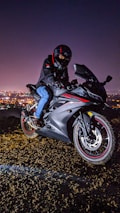 A person wearing a helmet and black jacket rides a sleek black motorcycle with red accents. The backdrop is a cityscape illuminated by night lights, creating a dynamic and adventurous atmosphere.