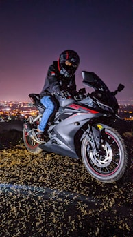 A person wearing a helmet and black jacket rides a sleek black motorcycle with red accents. The backdrop is a cityscape illuminated by night lights, creating a dynamic and adventurous atmosphere.