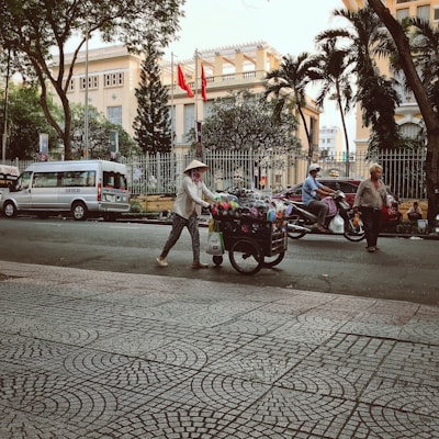 A street scene with a woman pushing a cart filled with various colorful goods. She is wearing a traditional conical hat and patterned clothing. The background includes a large building behind a fence, with several red flags flying. There are trees lining the street, a parked van, and motorcycles in motion. Pedestrians and motorcyclists are part of the busy urban environment.