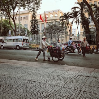 A street scene with a woman pushing a cart filled with various colorful goods. She is wearing a traditional conical hat and patterned clothing. The background includes a large building behind a fence, with several red flags flying. There are trees lining the street, a parked van, and motorcycles in motion. Pedestrians and motorcyclists are part of the busy urban environment.