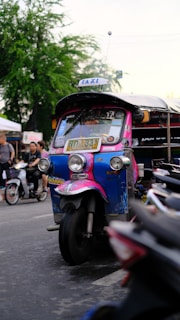 A colorful tuk-tuk with bright pink and blue detailing is parked on a bustling street, surrounded by other vehicles and pedestrians. Nearby, people are walking and engaging in activities typical of a busy urban area. Trees and outdoor market stalls can be seen in the background, adding to the vibrant city atmosphere.