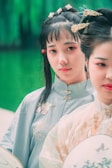 Two women wearing traditional clothing with intricate embroidery and ornaments in their hair are captured against a blurred green background. The woman in the foreground has a serene expression.