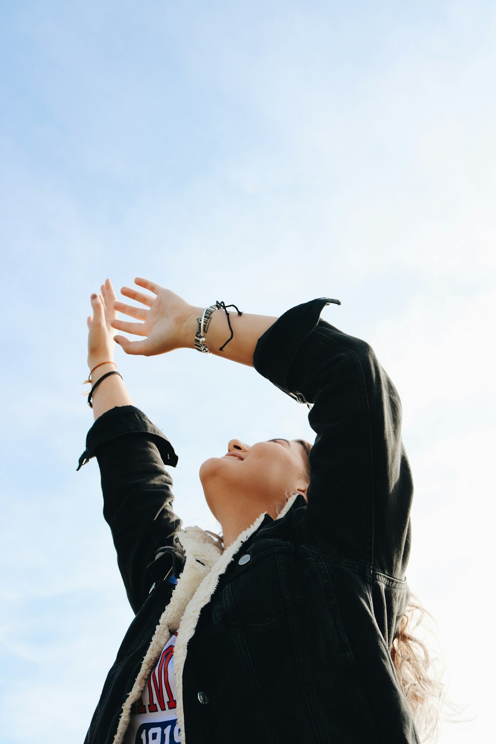 smiling woman wearing black and white jacket raising both hands while looking up under white and blue sky during daytime