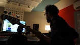 A person with a beard is silhouetted against a computer screen in a dimly lit recording studio. The person is holding a bass guitar and appears to be focused on a mixing console in front of them. The room features acoustic panels and a modern design, with a warm lamp softly illuminating the space.