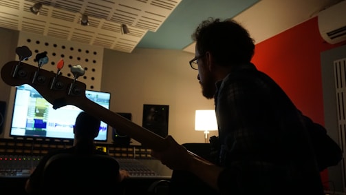 A person with a beard is silhouetted against a computer screen in a dimly lit recording studio. The person is holding a bass guitar and appears to be focused on a mixing console in front of them. The room features acoustic panels and a modern design, with a warm lamp softly illuminating the space.