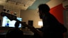 A person with a beard is silhouetted against a computer screen in a dimly lit recording studio. The person is holding a bass guitar and appears to be focused on a mixing console in front of them. The room features acoustic panels and a modern design, with a warm lamp softly illuminating the space.