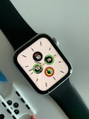 A modern smartwatch with a black strap displaying a digital clock face with multiple circular indicators. The screen shows the date as 'JEU 10' and various activity tracking metrics. The watch is placed on a light-colored surface with additional watch straps partially visible underneath.