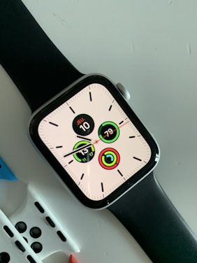 A modern smartwatch with a black strap displaying a digital clock face with multiple circular indicators. The screen shows the date as 'JEU 10' and various activity tracking metrics. The watch is placed on a light-colored surface with additional watch straps partially visible underneath.