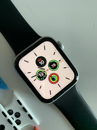 A modern smartwatch with a black strap displaying a digital clock face with multiple circular indicators. The screen shows the date as 'JEU 10' and various activity tracking metrics. The watch is placed on a light-colored surface with additional watch straps partially visible underneath.