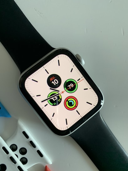 A modern smartwatch with a black strap displaying a digital clock face with multiple circular indicators. The screen shows the date as 'JEU 10' and various activity tracking metrics. The watch is placed on a light-colored surface with additional watch straps partially visible underneath.
