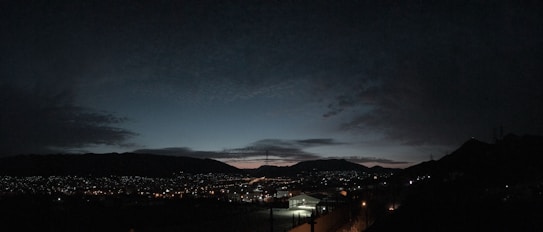 A panoramic view of a cityscape at dusk, with a dark sky overhead. Distant mountains outline the horizon, while scattered city lights create a sparkling effect across the landscape. The sky features a gradient from deep blue to lighter tones near the horizon, with wispy clouds.