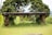 A rustic bench made of stone with a weathered appearance is set amidst lush greenery. It is supported by two stone pillars, surrounded by grass and vegetation, with trees in the background. The setting appears calm and natural, suggesting an outdoor garden or park area.