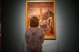 A person stands in front of a large, framed painting displayed in a museum or gallery. The painting depicts a religious or historical scene with several figures gathered around a large wooden cross in the background. The individual observing the artwork has a backpack and is facing the painting.