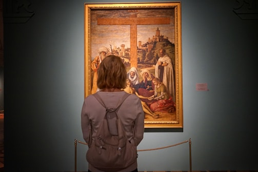 A person stands in front of a large, framed painting displayed in a museum or gallery. The painting depicts a religious or historical scene with several figures gathered around a large wooden cross in the background. The individual observing the artwork has a backpack and is facing the painting.