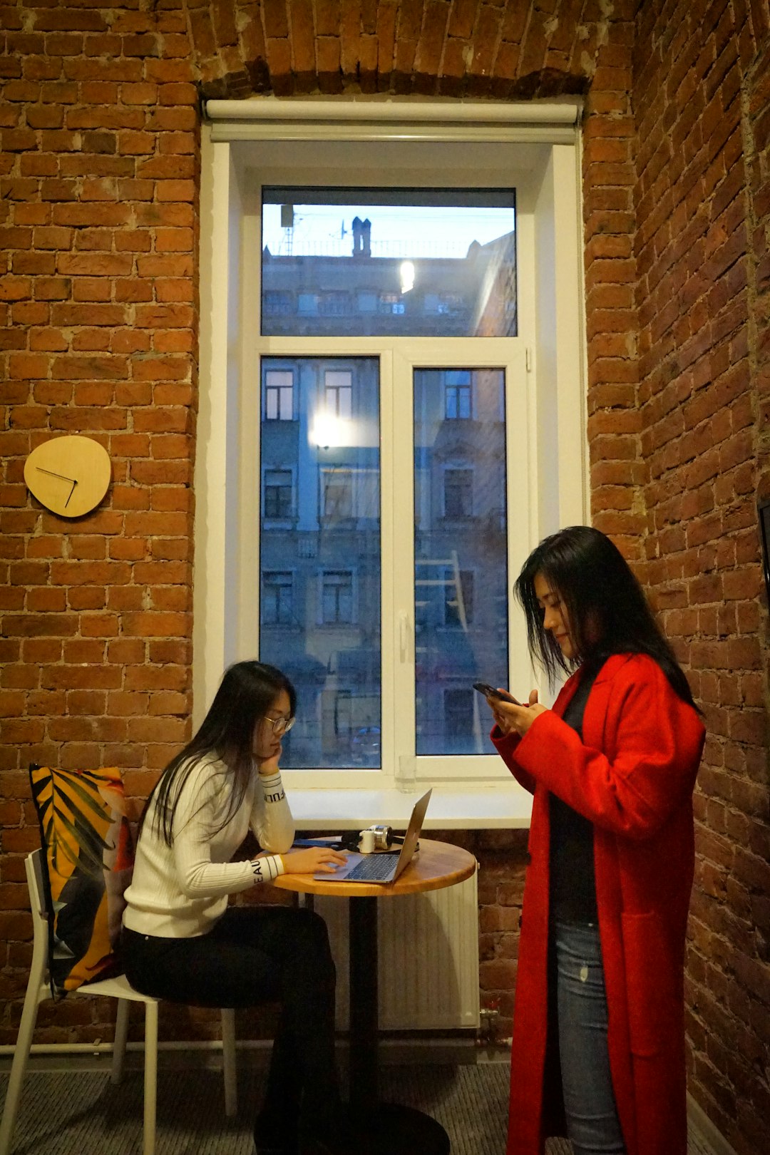 woman with red coat stands near woman at the table near window