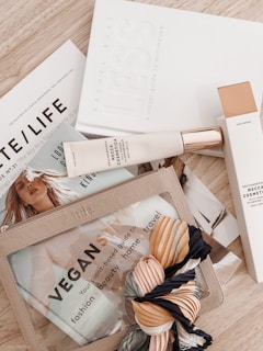 A flat lay image displaying a combination of lifestyle items including books, cosmetic products, and a colorful scarf. The books have minimalist and fashion themes, while the cosmetics feature elegant packaging. A transparent pouch contains a brochure titled 'VEGAN STYLE' that covers topics such as fashion and beauty.
